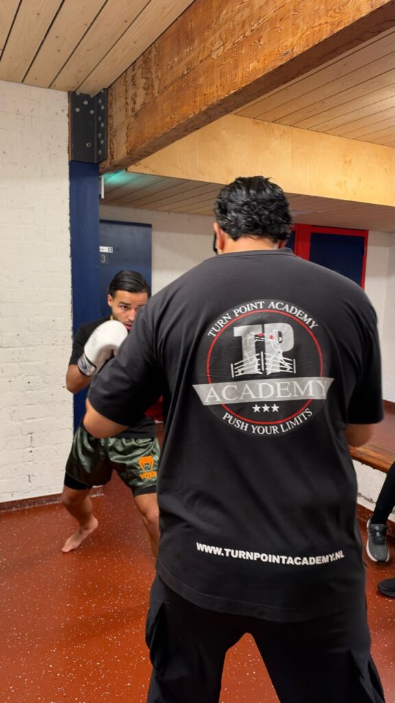 Een coach in een TPA-shirt begeleidt een kickbokser met witte bokshandschoenen.